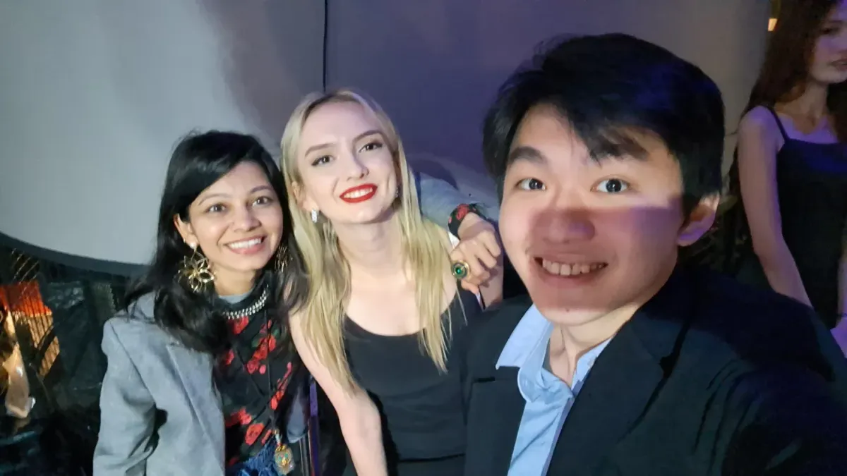 Selfie of a young man smiling in a dark jacket with two smiling women beside him, one with long blonde hair and one in a grey blazer.